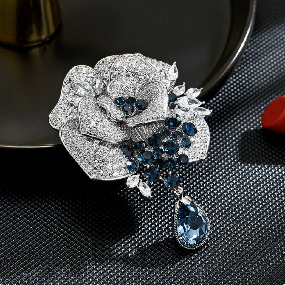 Front view of a silver-tone layered rose brooch with blue accents and a faceted teardrop dangle
