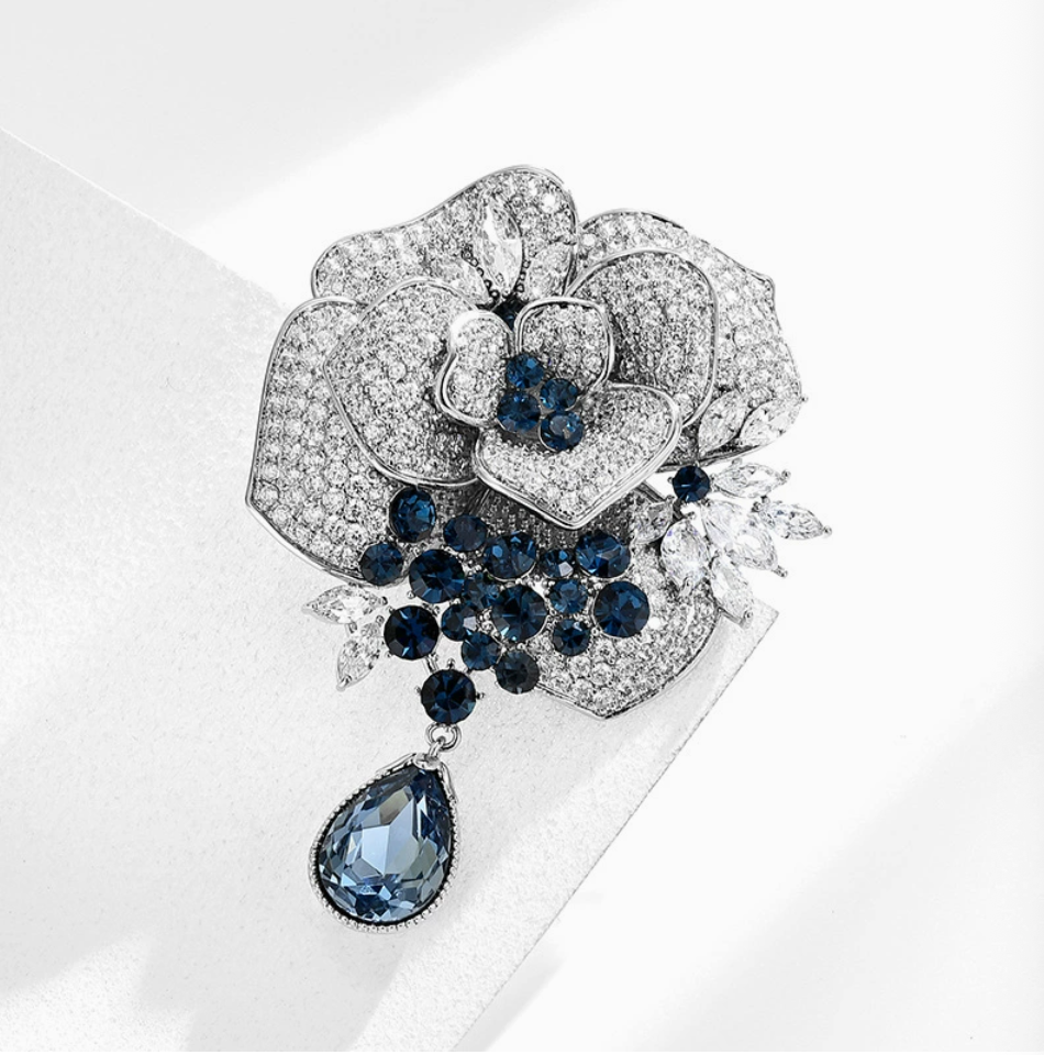 Silver-tone rose brooch with clear stones, dark blue accents and a blue teardrop dangle on a white background