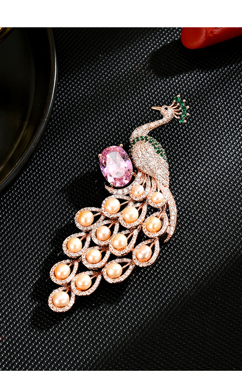 Rose-gold-tone peacock brooch with pink and green stone accents and freshwater pearls on a black textured surface