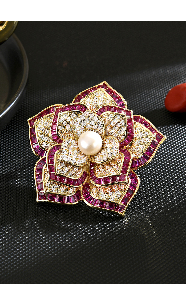 Fuchsia and gold-tone camellia brooch with cubic zirconia and a freshwater pearl center on a black textured surface